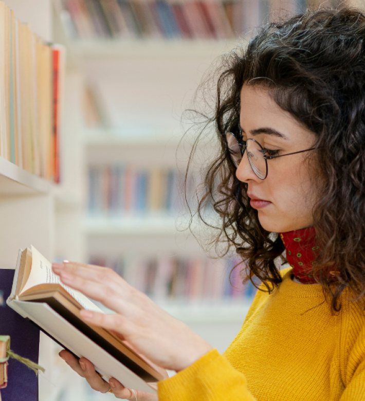 En kvinde med skulderlangt mørkt krøllet hår og briller, iført en gul sweater og rødt tørklæde om halsen. Står og kigger i en bog ved en lys bogreol